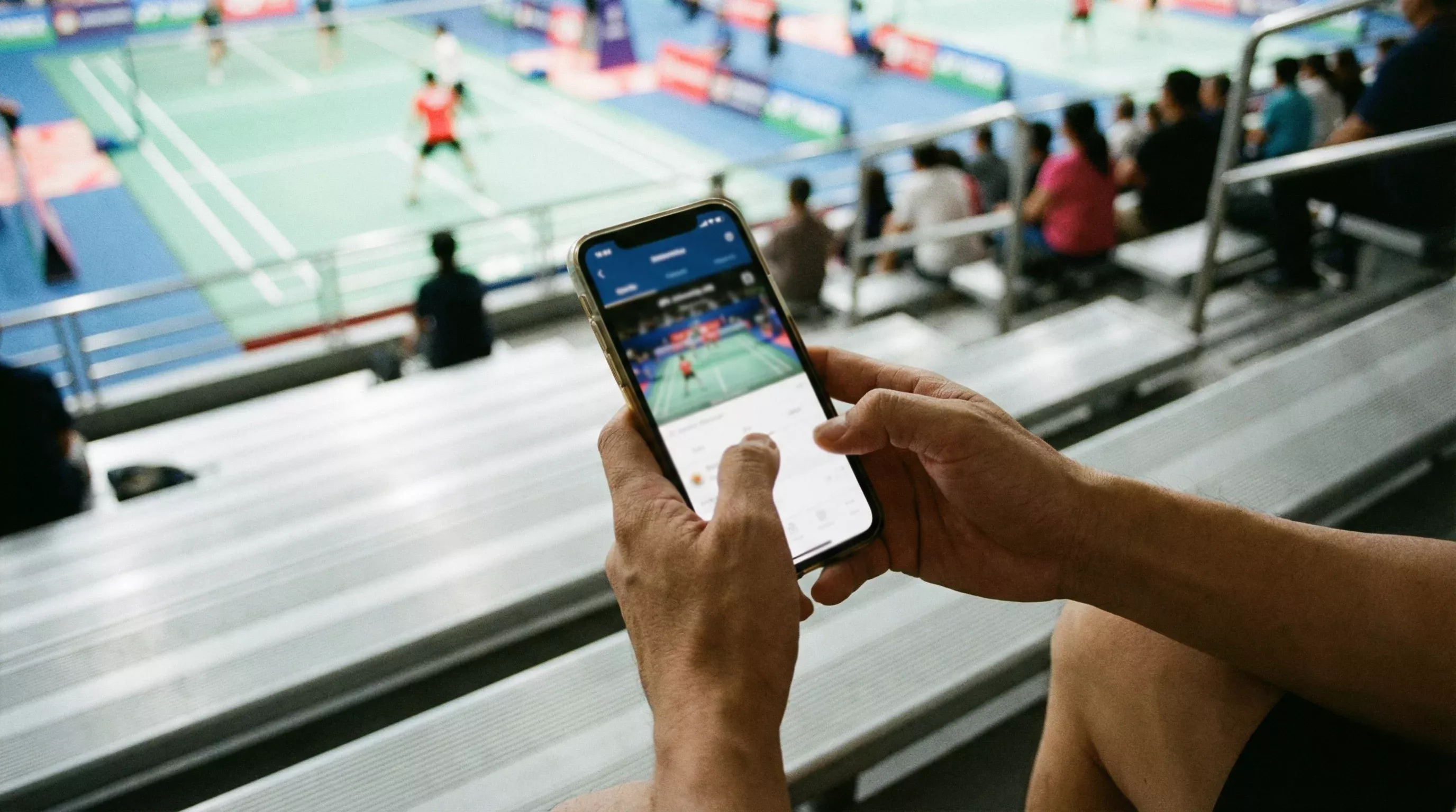 Person hält Smartphone und beobachtet ein Badminton-Spiel im Hintergrund